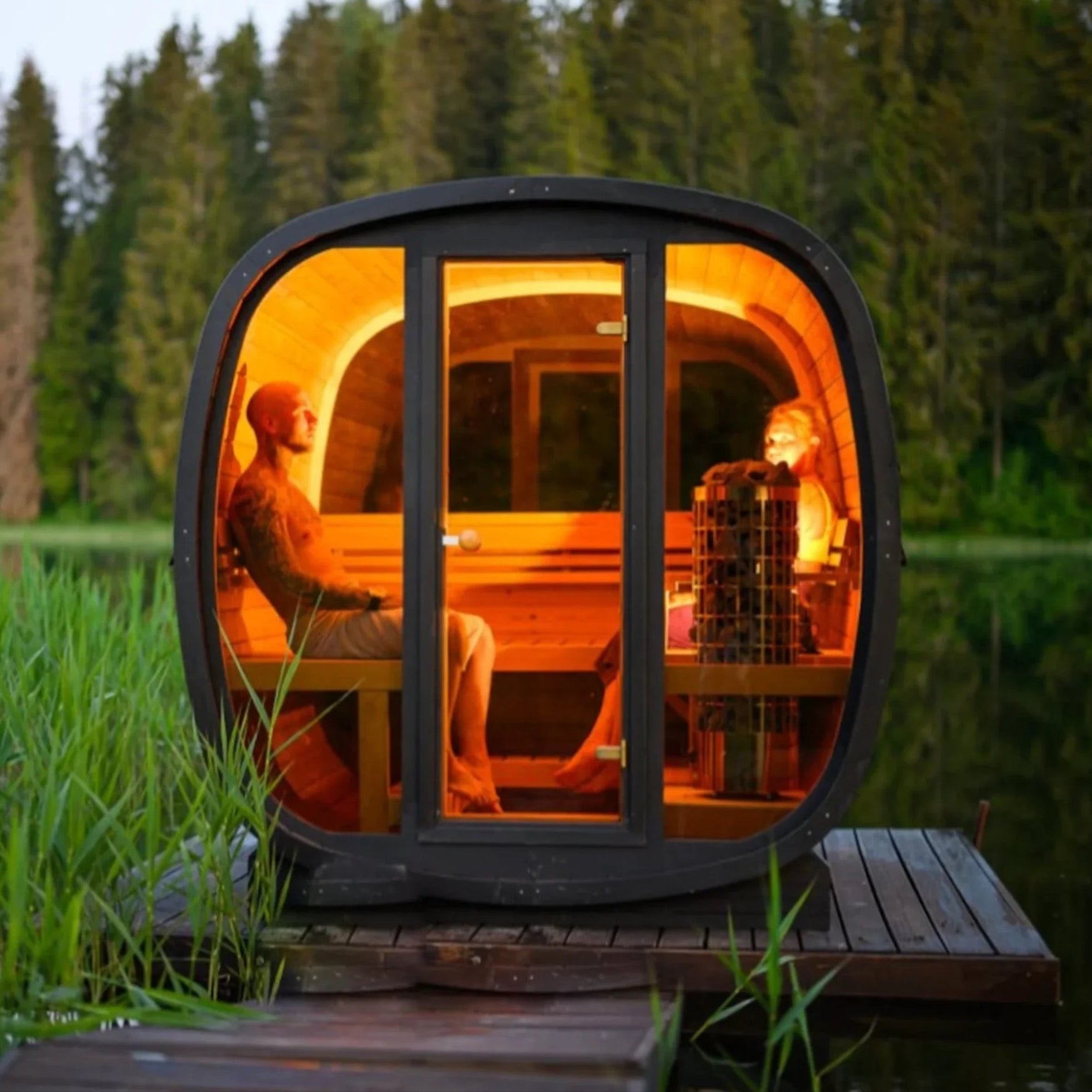 Luxury outdoor barrel sauna with a panoramic glass viewing window on a lake dock at sunset.