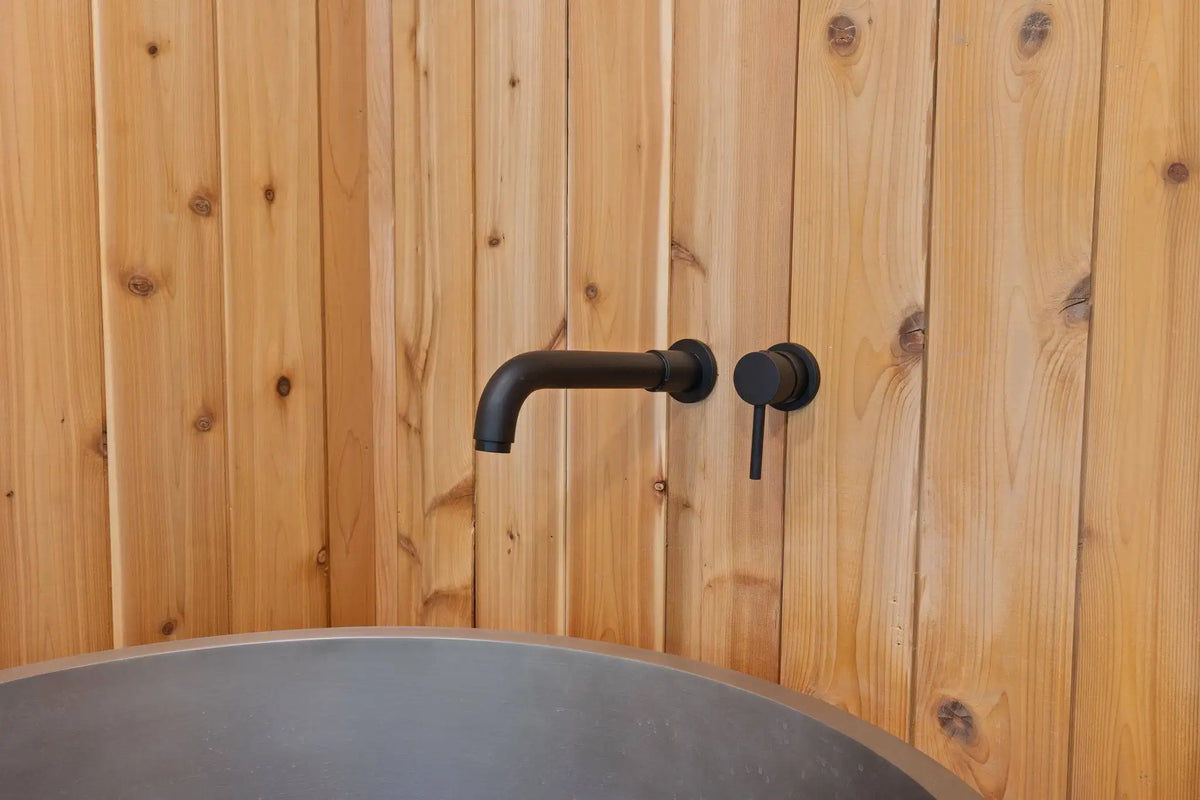 A matte black wall-mounted faucet and matching control handle installed on a light natural wood plank wall above a dark gray round basin.