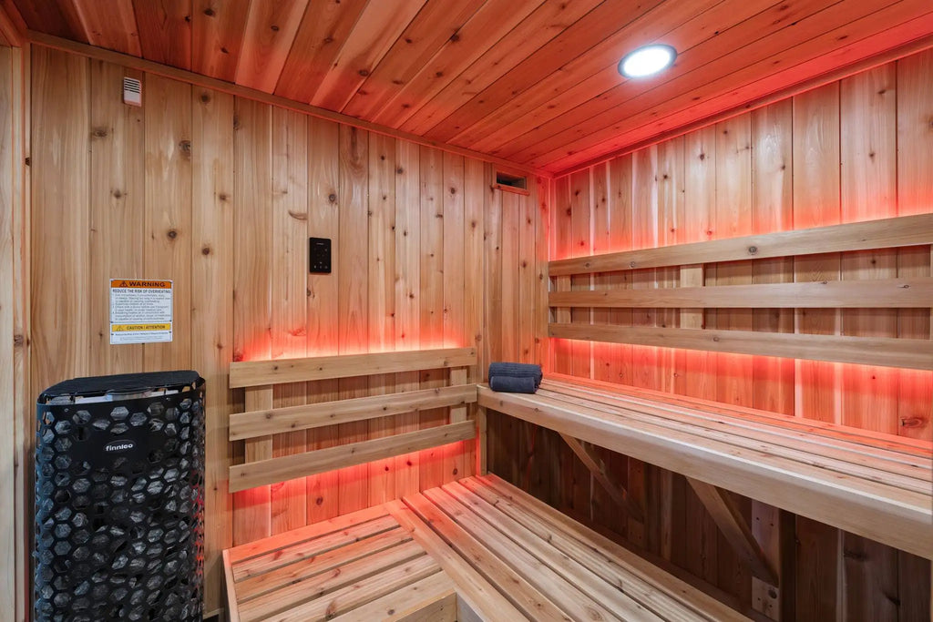 A wooden sauna interior with tiered wooden benches, wood-paneled walls and ceiling, red ambient lighting, a black stone heater, and a small towel on one bench.
