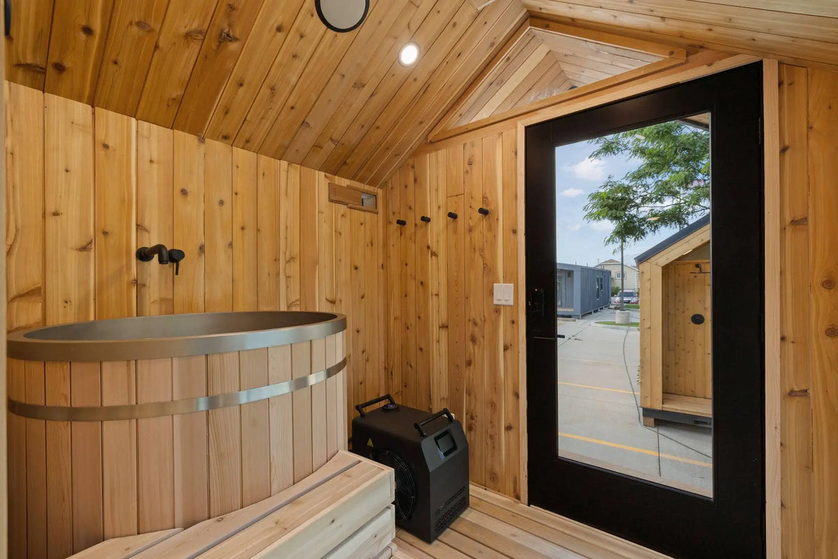 A wooden sauna interior with a round wooden hot tub, wooden benches, wood-paneled walls and ceiling, a black-framed glass door, and a small black equipment box on the floor.