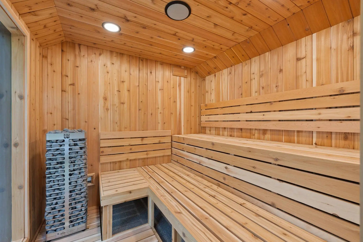 A wooden sauna interior with tiered wooden benches, wood-paneled walls and ceiling, recessed ceiling lights, and a stack of gray sauna stones on the left.