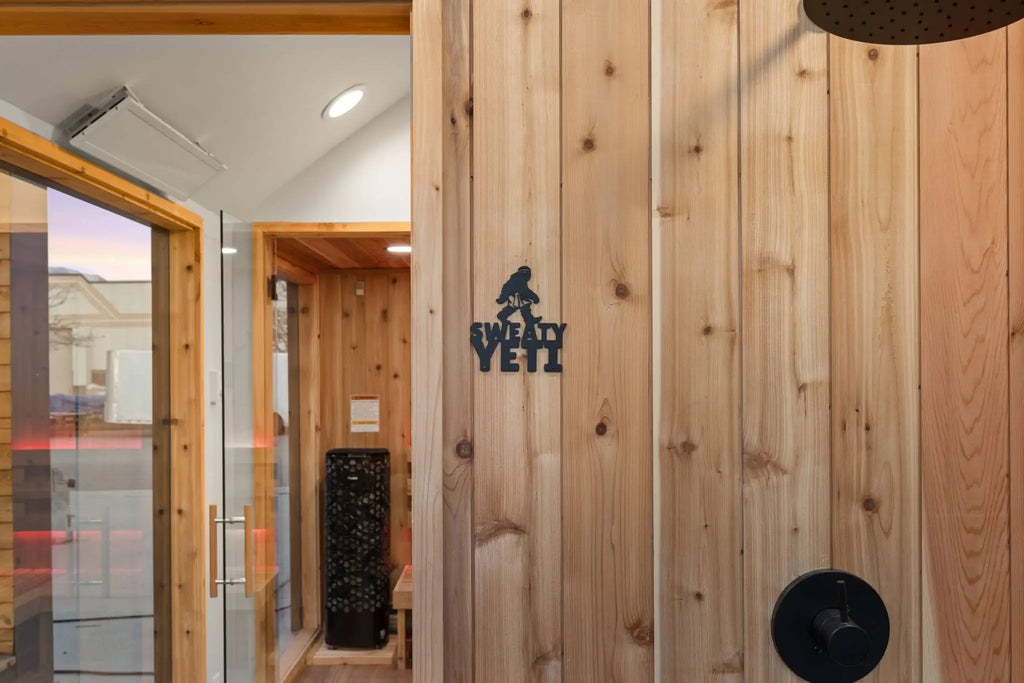 Wood-paneled sauna door with a black metal 'Sweaty Yeti' logo, glass entryway, and indoor sauna room visible in the background.