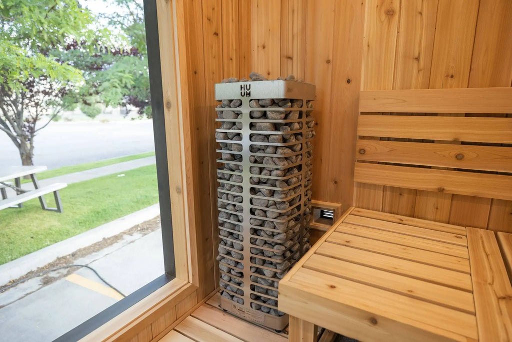 A wood-burning sauna interior with a tall metal log stack heater, wooden benches, and a large window showing an outdoor lawn and trees.
