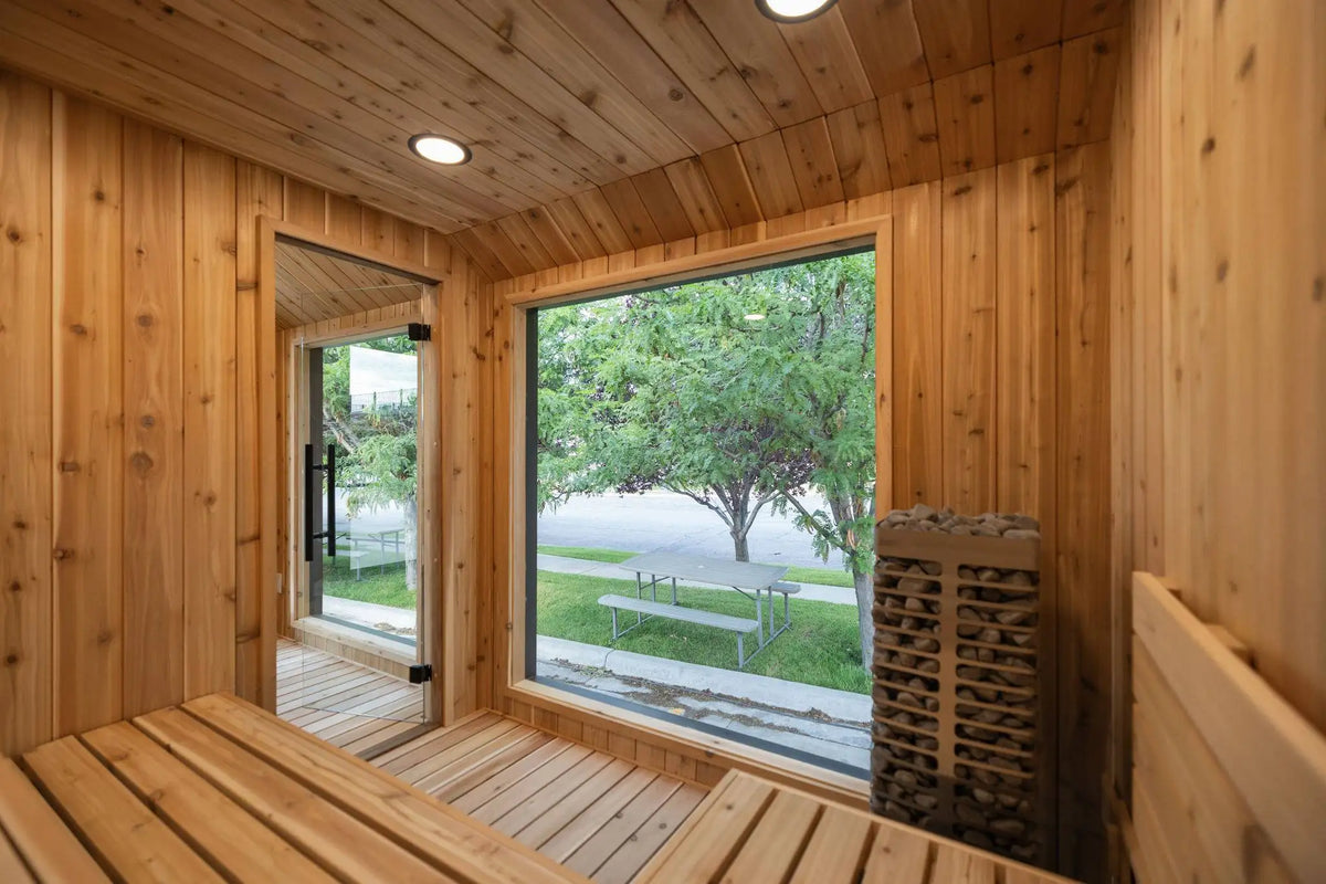 A wooden sauna interior with a wood-bench seating area, paneled walls and ceiling, a large window showing an outdoor green lawn and trees, a glass door, and a wood-burning stove stack on the right.