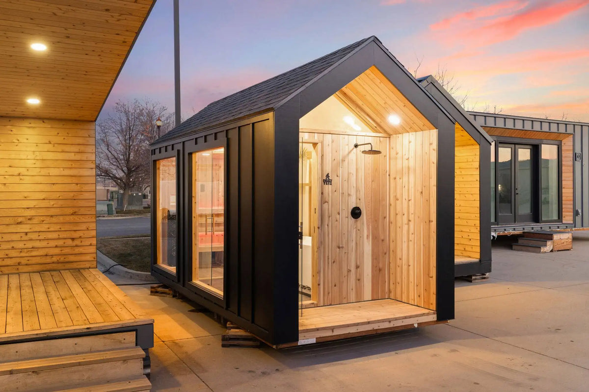 A modern small cabin or sauna-style outdoor room with a black metal frame, wood-paneled walls, large glass windows, and an open front porch, shown at dusk beside other similar structures.