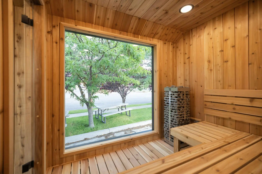 A wooden sauna interior with tiered benches, a large window showing green trees outside, and a stacked wood fire basket.