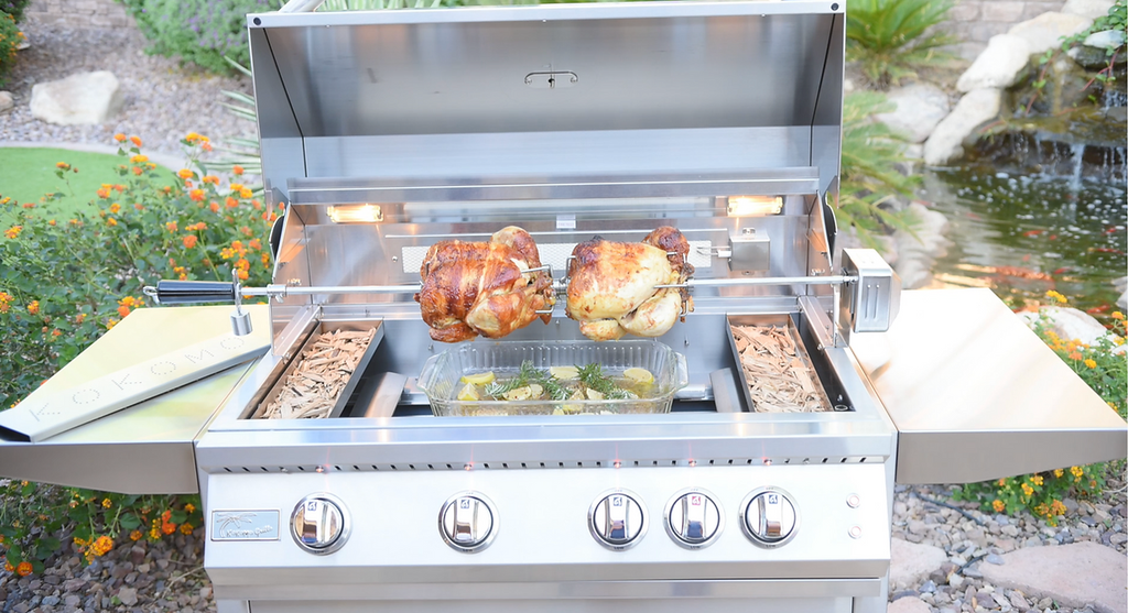 Stainless steel grill with two roasted chickens on a patio with a garden and water feature in the background.