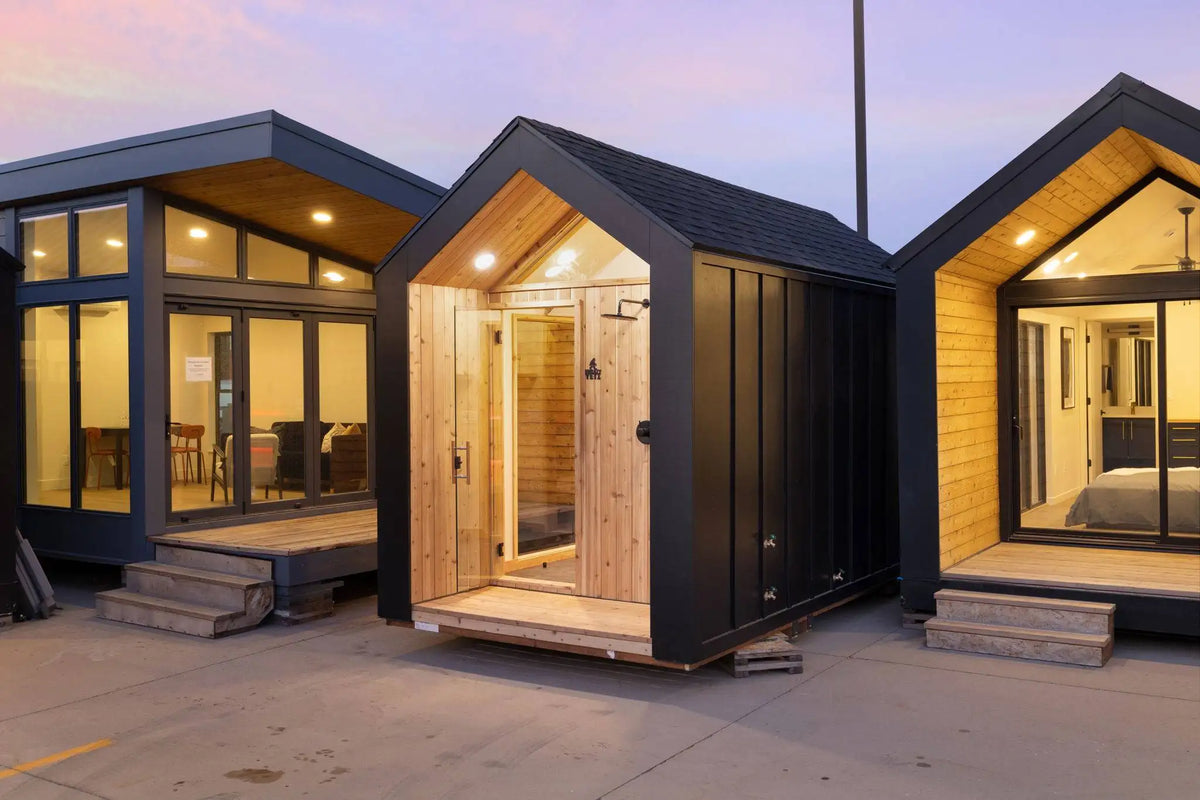 Three modern modular sauna units with black metal siding and wood interiors, shown side by side at dusk with the center unit open to reveal a sauna interior.