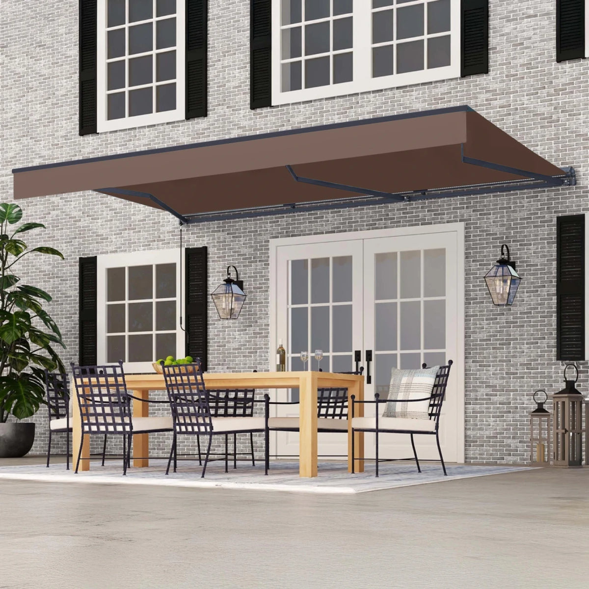 A brown retractable patio awning extends over a dining area on a house porch. The scene shows a gray brick home exterior with white-framed windows, black shutters, outdoor lanterns, a wooden dining table, and black mesh chairs.