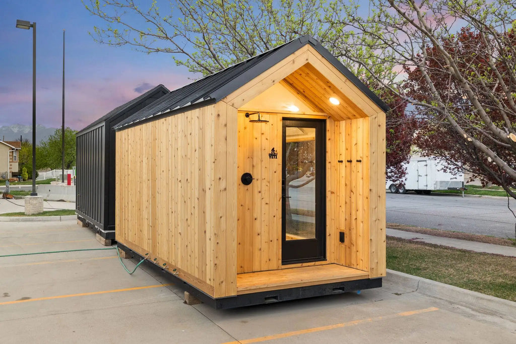 A small modular cabin home with light natural wood siding and a black metal roof, parked outdoors on a concrete pad with trees and a street visible in the background.