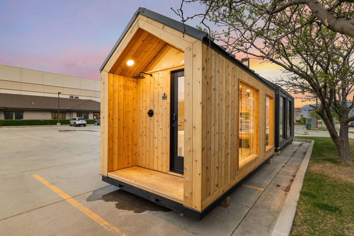 A modern modular tiny home with light natural wood siding and a dark roof, parked outdoors on a concrete pad near a tree at dusk.
