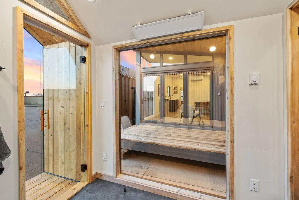 A modern interior sauna space with a large glass door and sliding glass panels framed in natural wood, opening onto a deck and an outdoor patio at dusk. The room has light-colored walls, a wood floor, and a white air conditioner mounted on the ceiling.