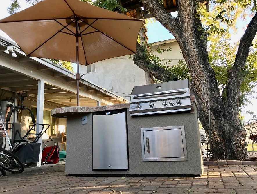Outdoor kitchen with grill and refrigerator under a large umbrella on a patio.