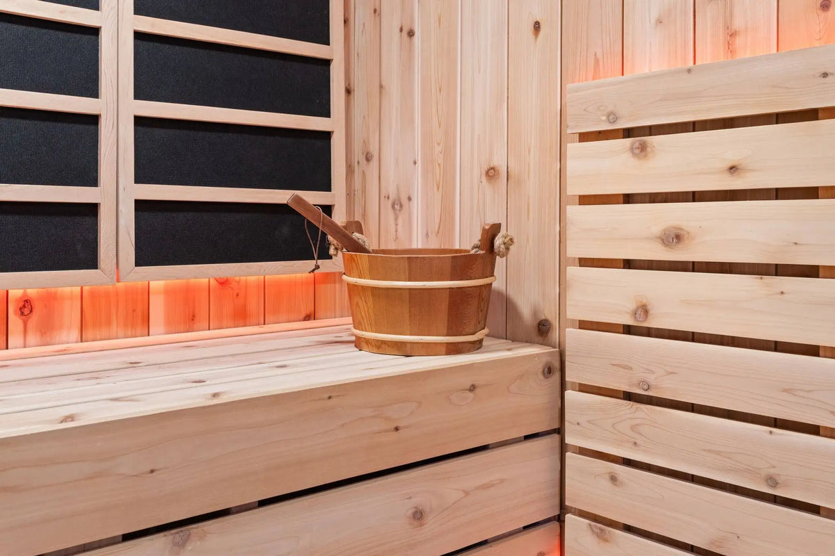 A traditional wooden sauna interior with light pine wood walls, a wooden bench, a wooden bucket, and a wooden ladle, illuminated by warm orange-red ambient lighting.