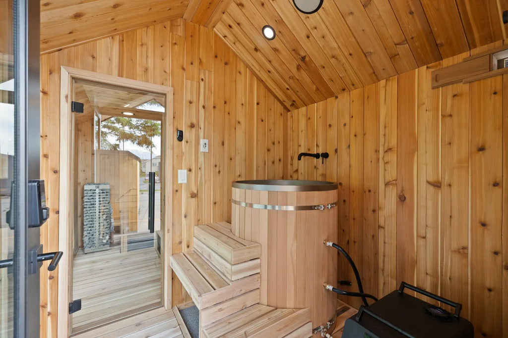 A rustic wooden sauna interior with light natural wood walls, a wooden bench, a round wooden hot tub, and a glass door opening to an outdoor deck.
