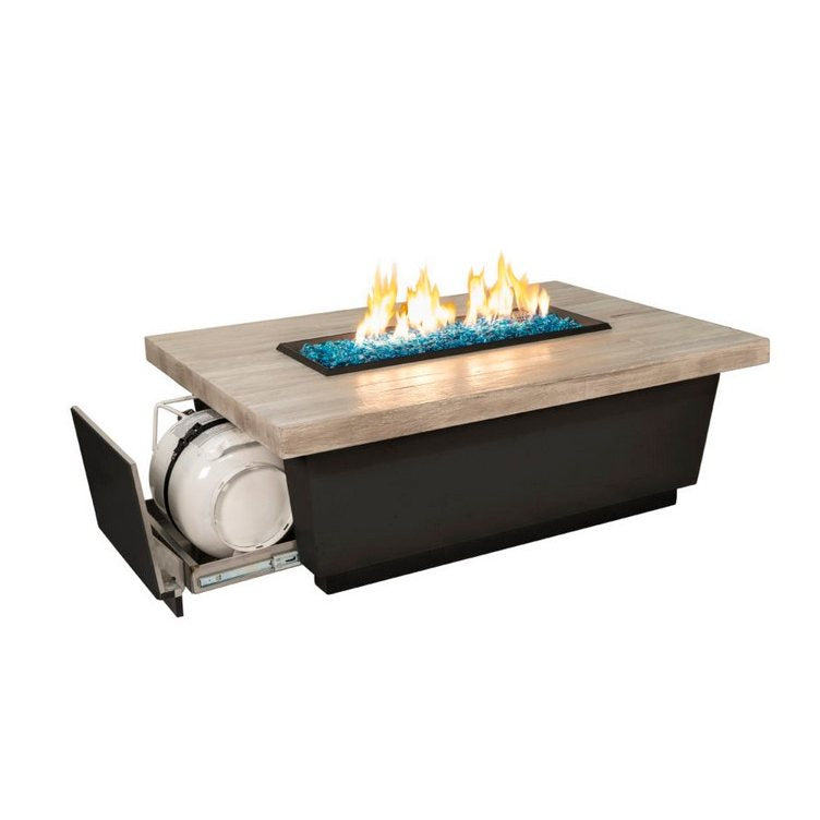 A rectangular outdoor fire pit table with a light wood-look top, black base, and blue glass stones with active flames. A pull-out storage compartment on the left holds a white propane tank, shown against a plain white background.