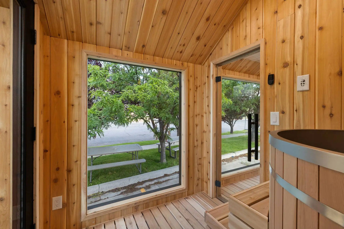 Wood-paneled interior room with a large window showing trees and a paved outdoor area, plus a wooden hot tub with metal bands visible on the right.