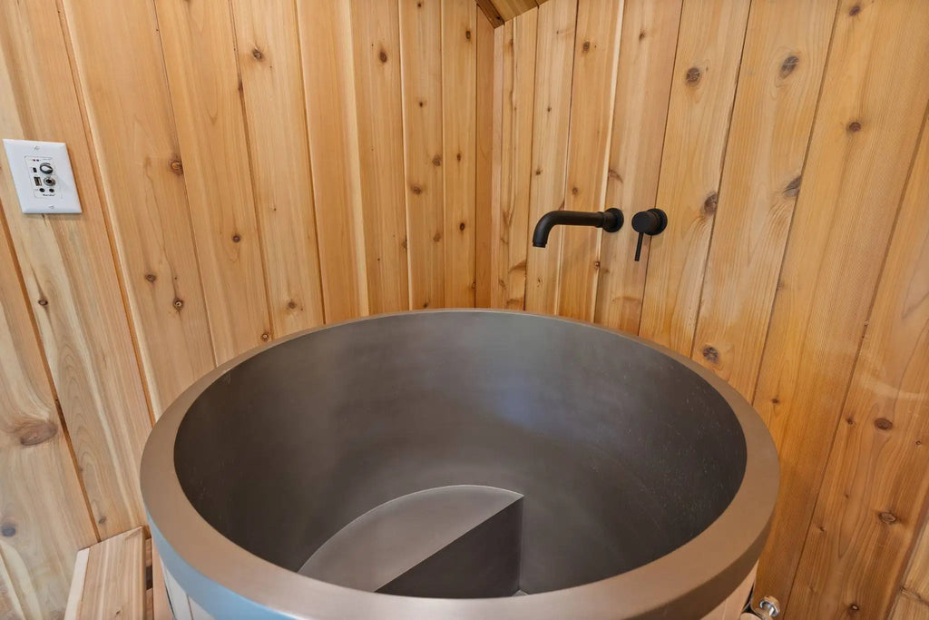 A large round matte gray concrete or stone vessel with a black wall-mounted faucet, set against a wood-paneled wall in a rustic interior.