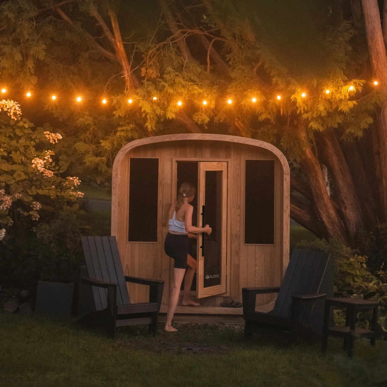 Person opening a wooden sauna door in a garden setting with string lights.