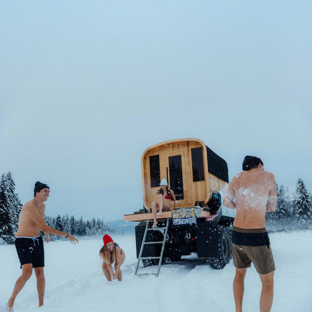 People in swimsuits standing around a small wooden cabin in the snow.