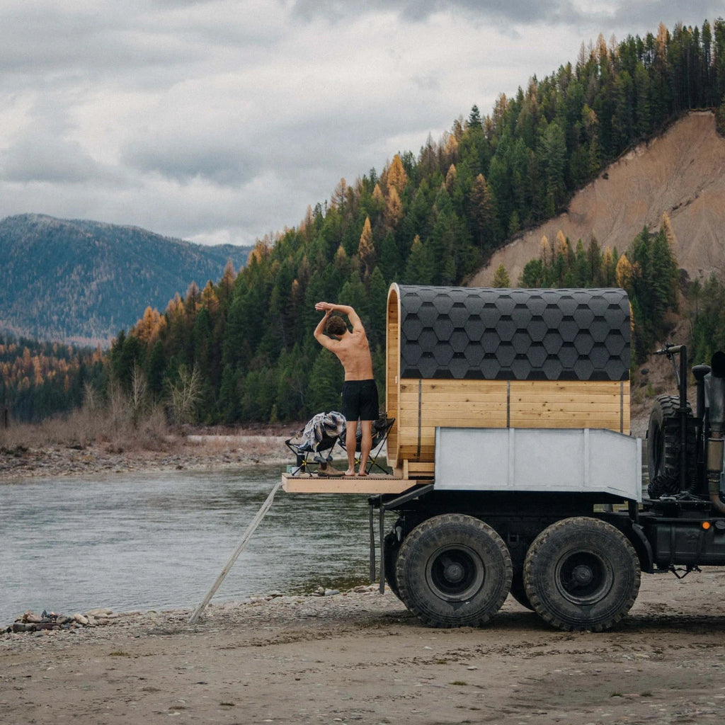 Person standing on a truck with a small wooden cabin on top, near a lake and forest.