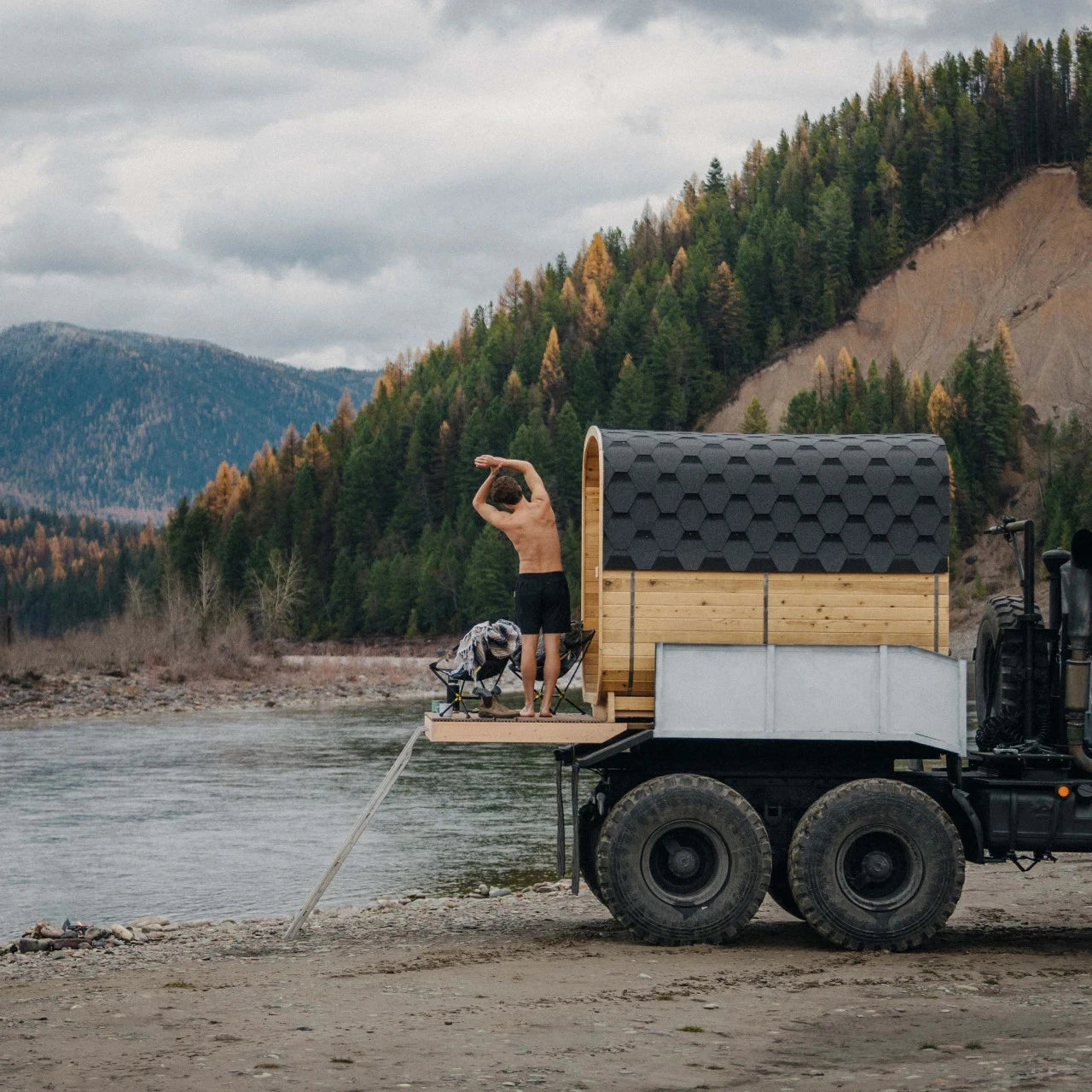 Person standing on a truck with a small wooden cabin on top, near a lake and forest.