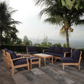 Outdoor patio teak furniture set navy cushions on a stone patio with palm trees in the background.