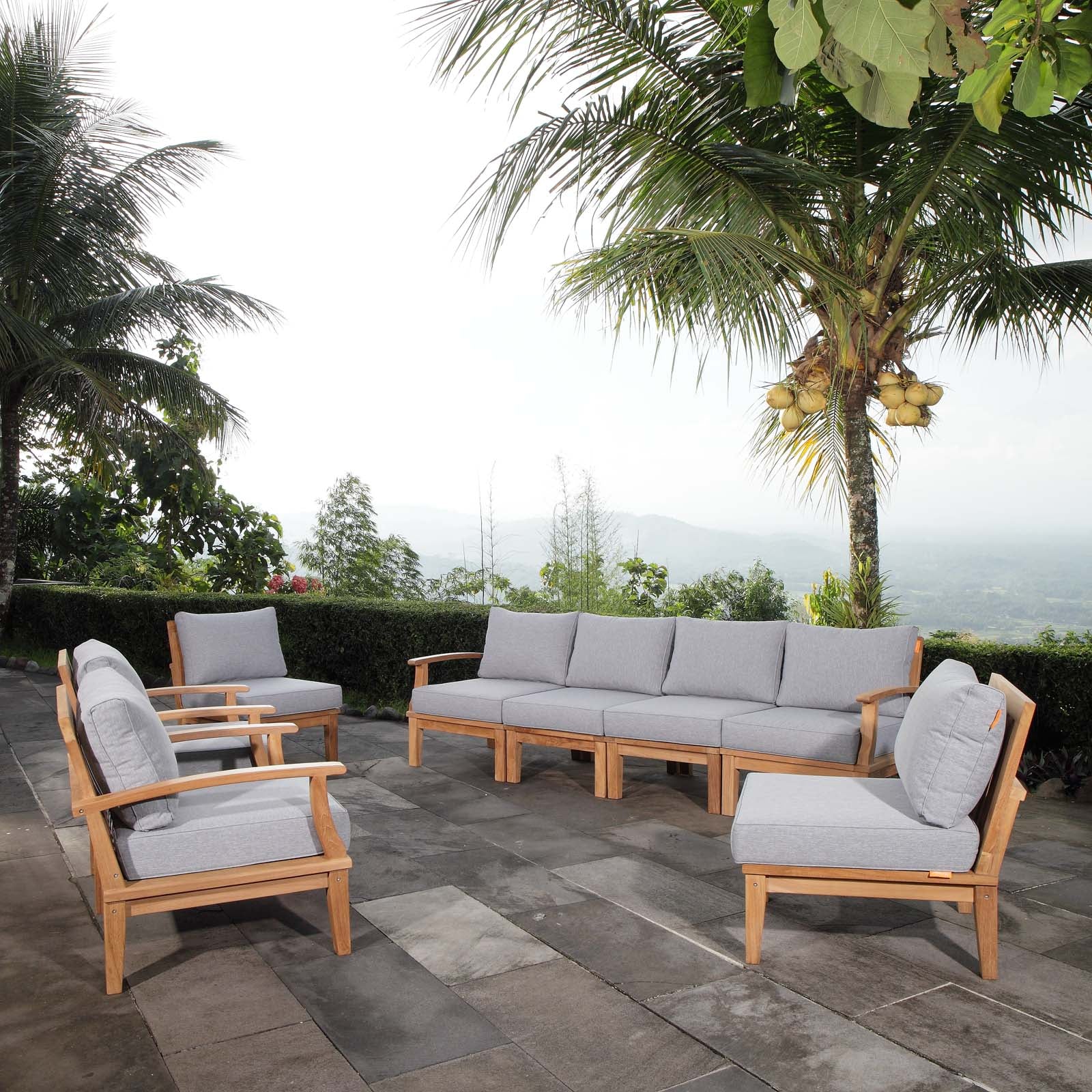 Outdoor patio furniture set with wooden frames and gray cushions on a stone patio with palm trees in the background.