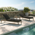 Two lounge chairs with black cushions by a poolside with a scenic background.