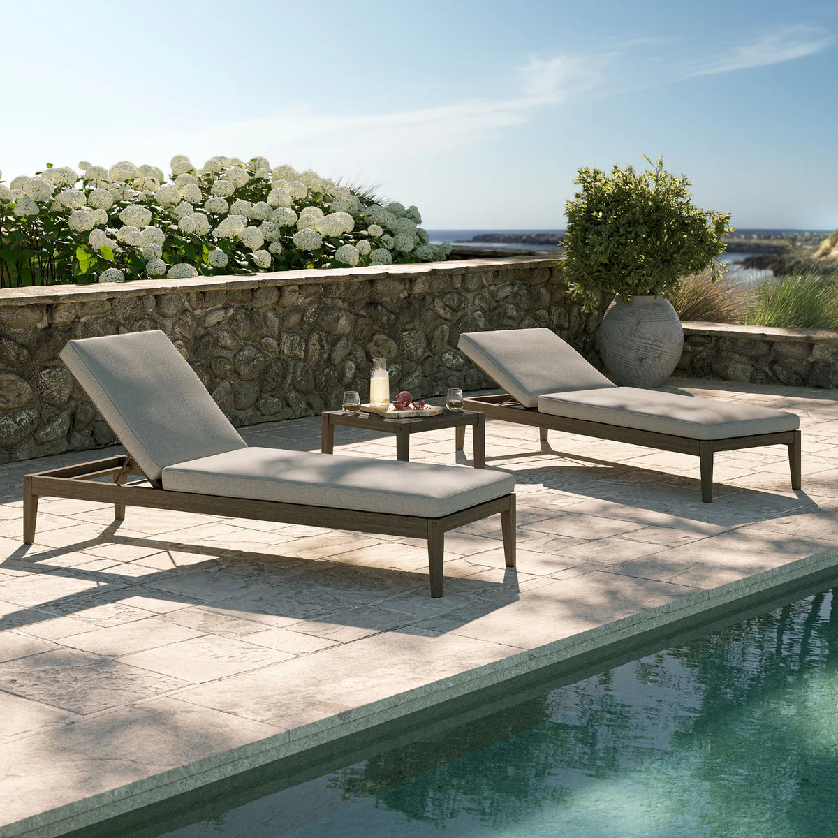 Two lounge chairs with white cushions by a poolside with a scenic background.
