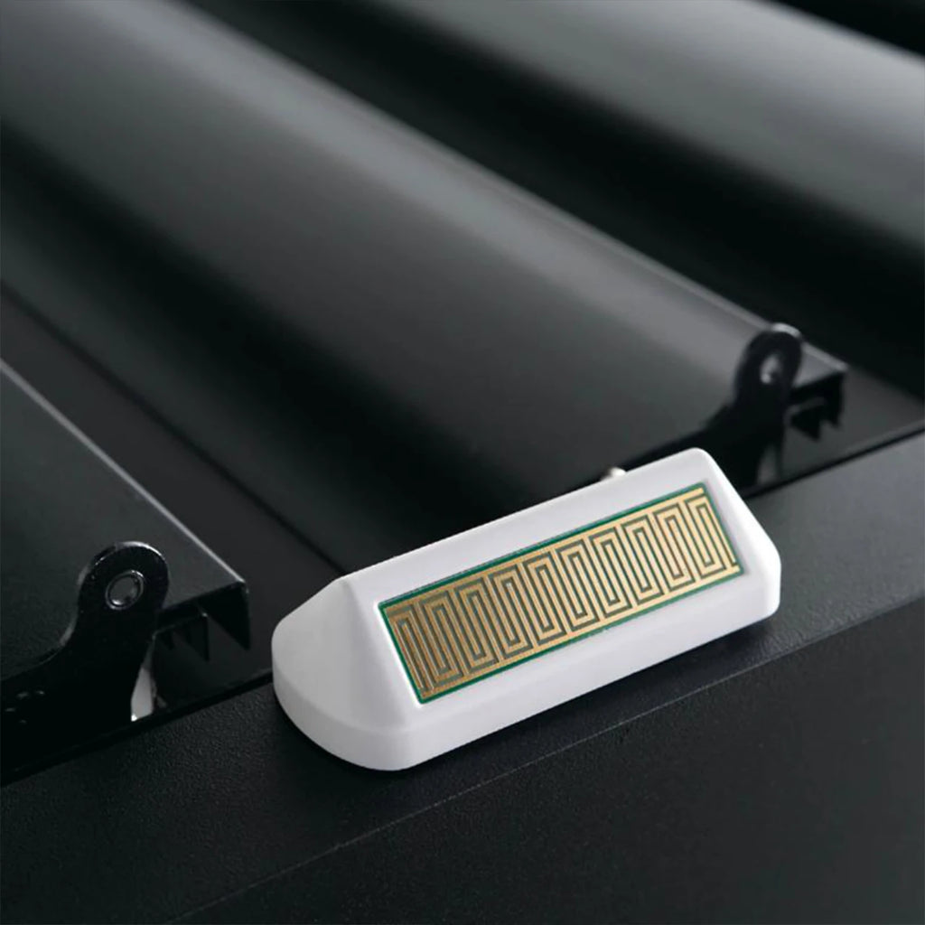 A close-up of a white rectangular electronic sensor or sensor module with a gold-colored rectangular circuit pattern on its top surface, mounted on a black metal surface.