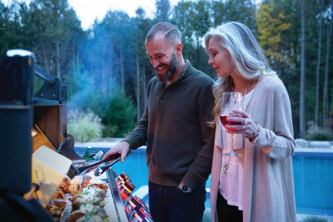 Man and woman grilling outdoors by a pool with trees in the background