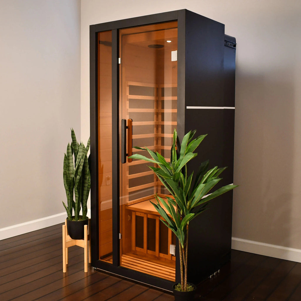 Infrared sauna with wooden interior and black exterior, surrounded by plants on a wooden floor.