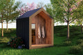A modern outdoor sauna structure with a dark charcoal side wall, warm wood-paneled front opening, and a peaked roof, shown in a grassy yard with trees and flowers.