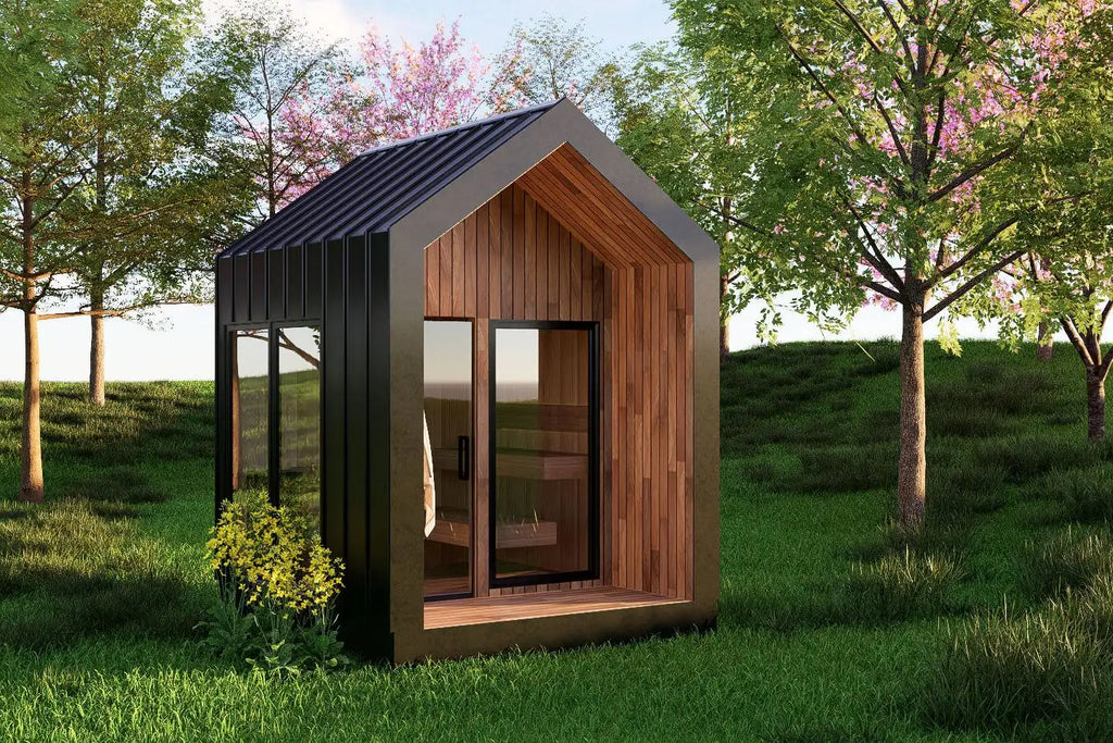 A modern sauna with a dark metal exterior, wood-paneled front, and large glass windows, shown standing on grass in a landscaped yard with trees.