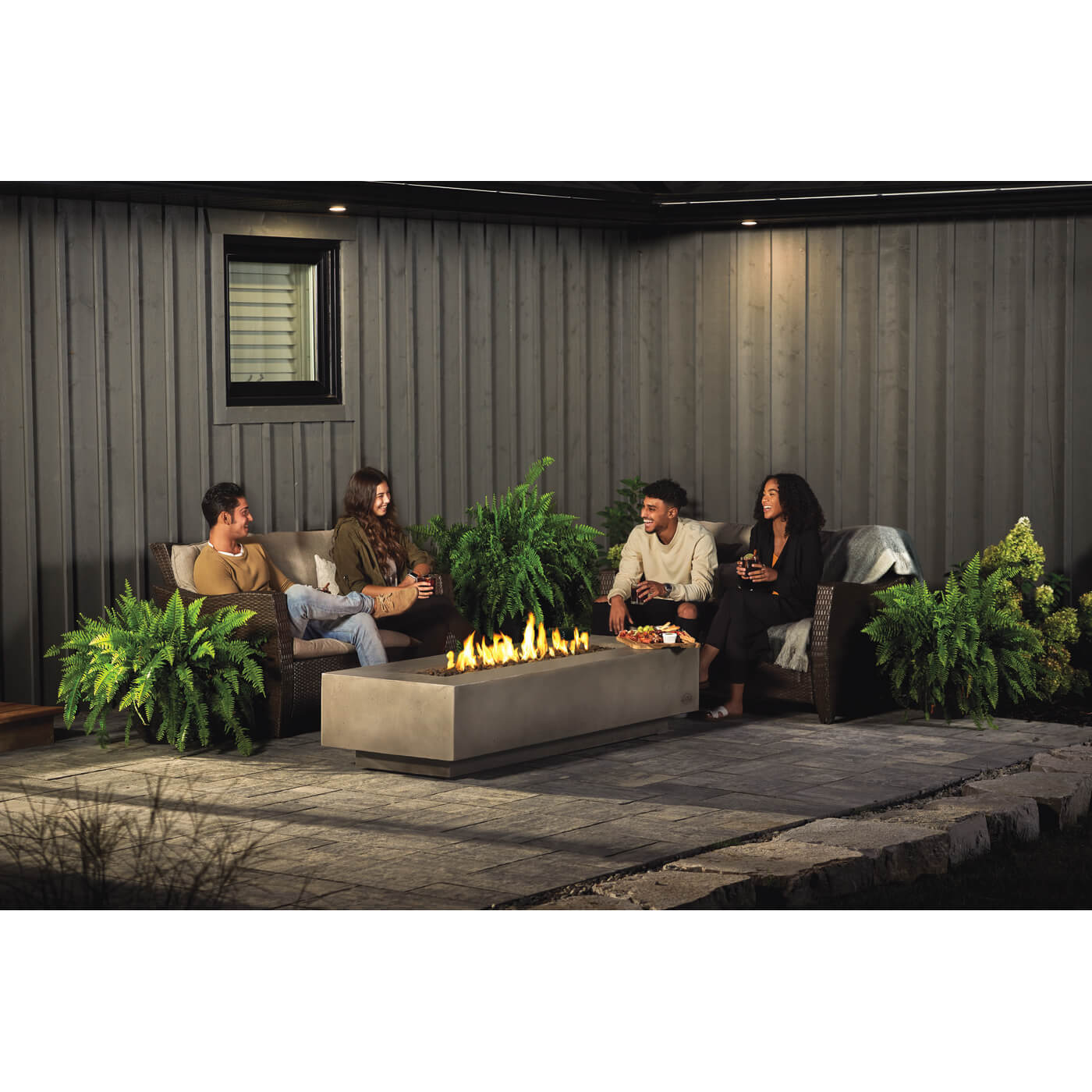 People sitting around a fire pit in an outdoor setting with plants and a wall.
