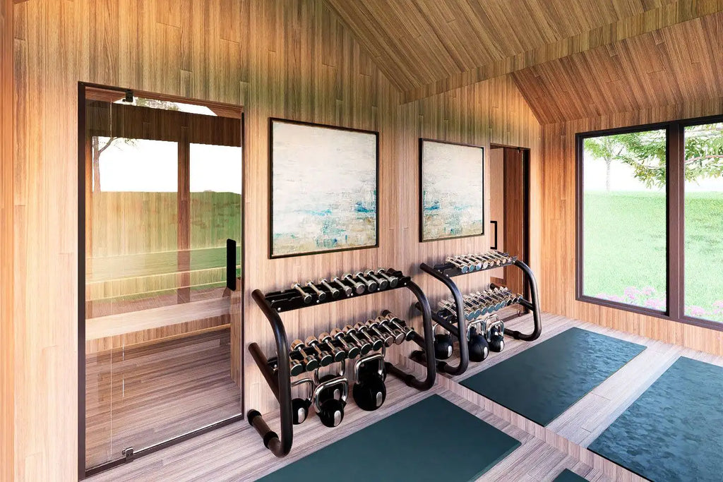 A modern indoor fitness room with wood-paneled walls and ceiling, featuring two black dumbbell racks, weight plates, dark green exercise mats, and large windows looking out onto a grassy lawn.