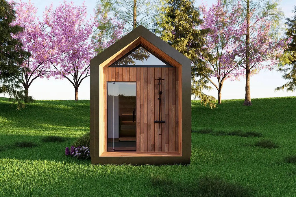 A modern wooden sauna cabin with a pitched roof, glass door, and visible shower fixture, set in a grassy outdoor scene with flowering trees in the background.