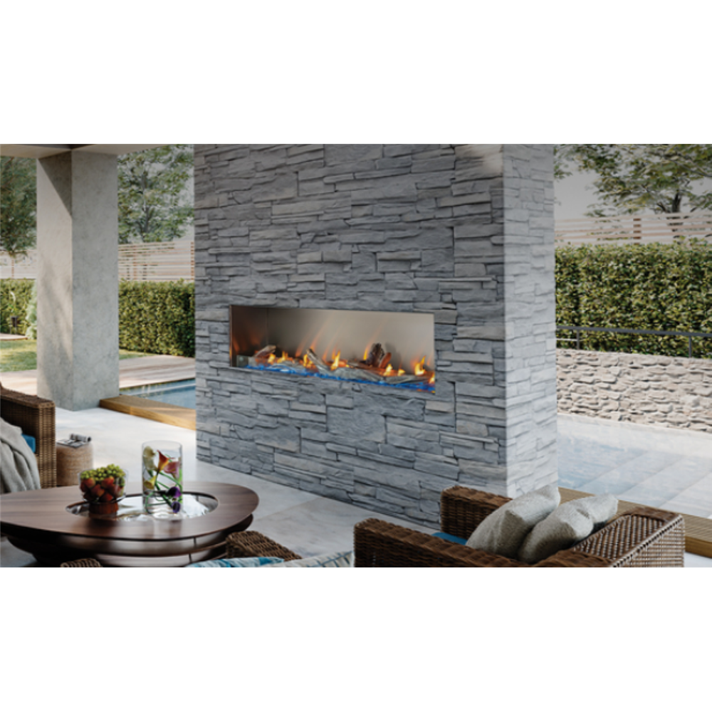 A modern linear fireplace built into a gray stone wall, shown on an outdoor patio with wicker furniture, a round coffee table, and greenery in the background.