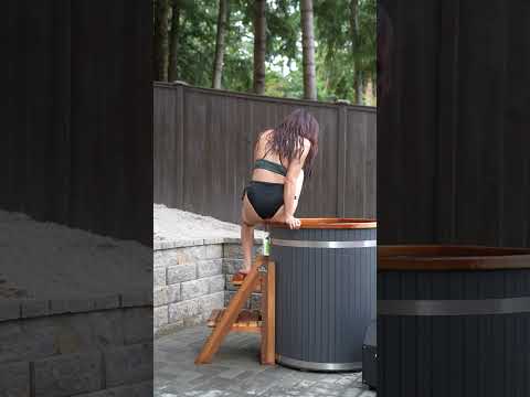 A woman in a black bikini stands on a wooden ladder next to a dark gray barrel col tub with a wooden rim, set outdoors on a stone patio with a wooden fence and trees in the background.