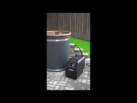 A black cold  tub and chiller with a hose connected, placed next to a gray hot tub barrel on a paved backyard patio with artificial grass and a wooden fence.