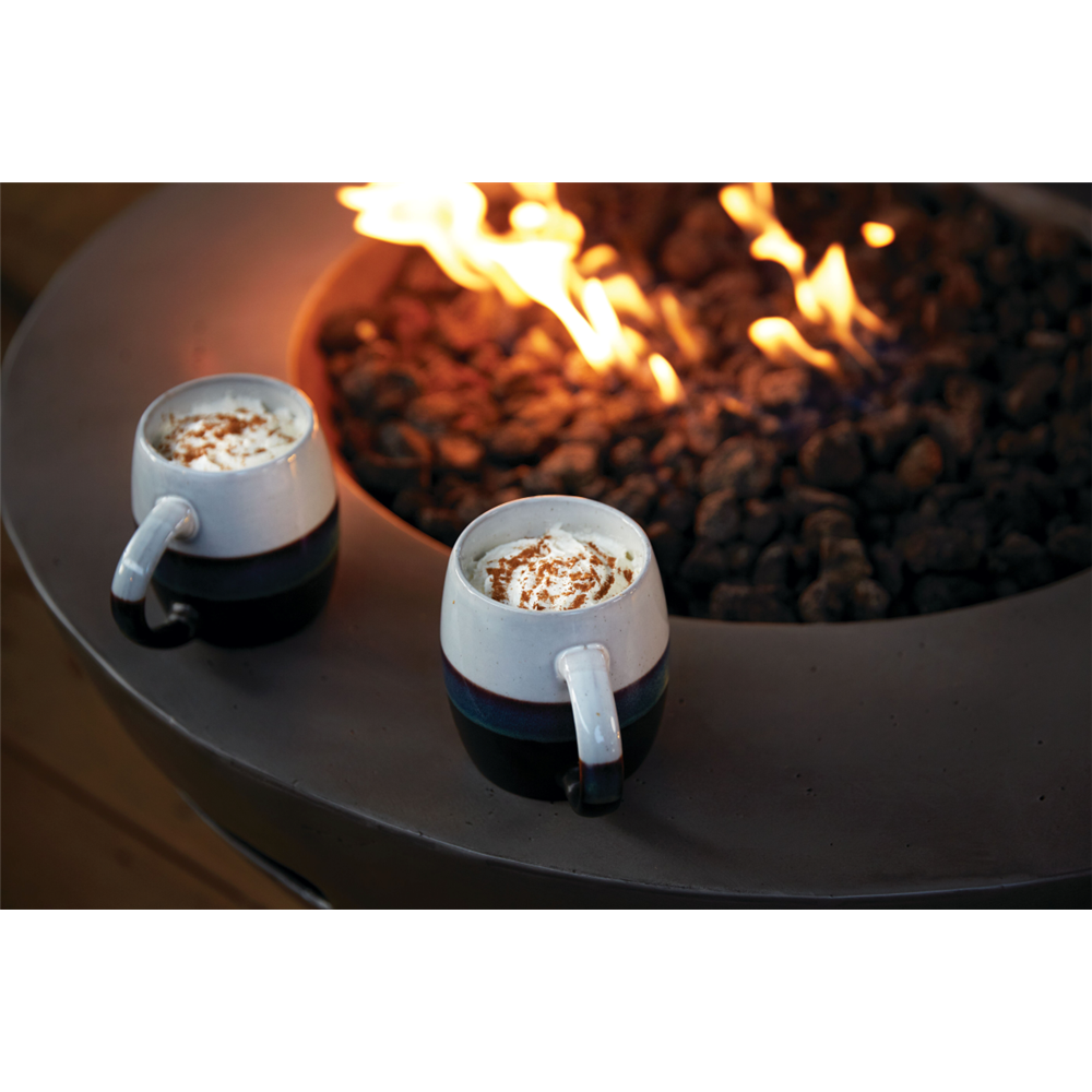 Two ceramic mugs filled with a hot drink and topped with whipped cream and cocoa powder, placed on the rim of a fire pit with flames burning in the background.