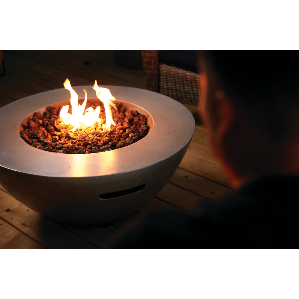 A round concrete fire pit table with a bowl-shaped design, filled with glowing orange and yellow flames over dark decorative stones, set on a wooden deck with a person's silhouette visible in the foreground.