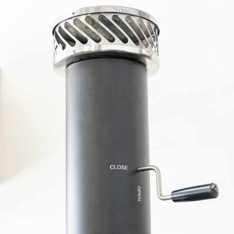 A close-up of a dark gray cylindrical chimney cap with a silver metal vent top and a hand crank handle labeled CLOSE and OPEN.