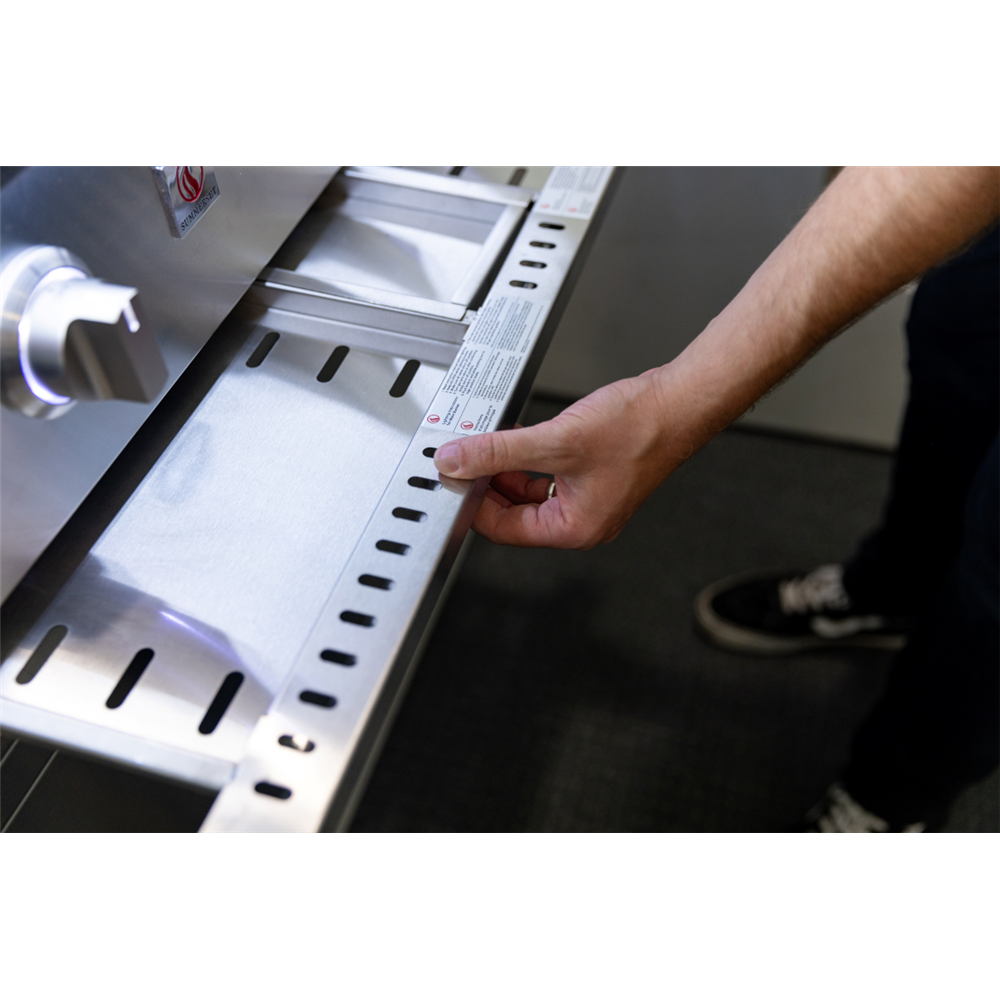 A close-up of a person's hand pointing to a measurement guide or instruction sheet attached to a stainless steel grill or smoker, with the person's arm and lower legs visible in a real-world setting.