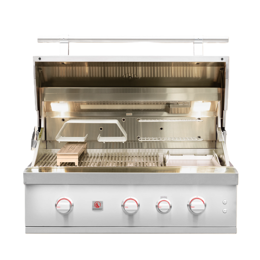 A stainless steel outdoor gas grill with its lid open, showing the lit interior grates and warming lights. The front features four control knobs with red markings and a small red logo badge.