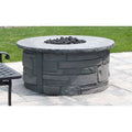 A round outdoor fire pit table made of gray faux-stone stacked blocks, with a flat stone-look top and black fire rocks inside, set on a light stone patio with green grass in the background.