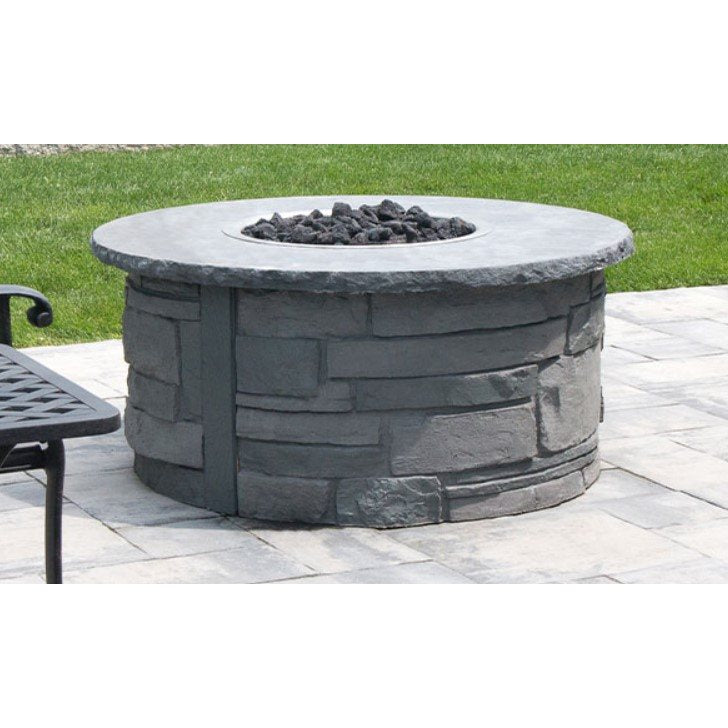 A round outdoor fire pit table made of gray faux-stone stacked blocks, with a flat stone-look top and black fire rocks inside, set on a light stone patio with green grass in the background.