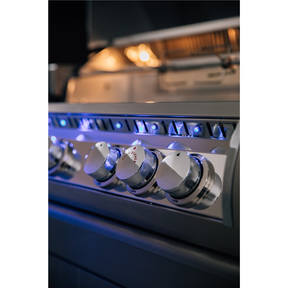 Close-up of a stainless steel outdoor grill control panel with four white rotary knobs and blue LED indicator lights, with the blurred grill body and flames in the background.