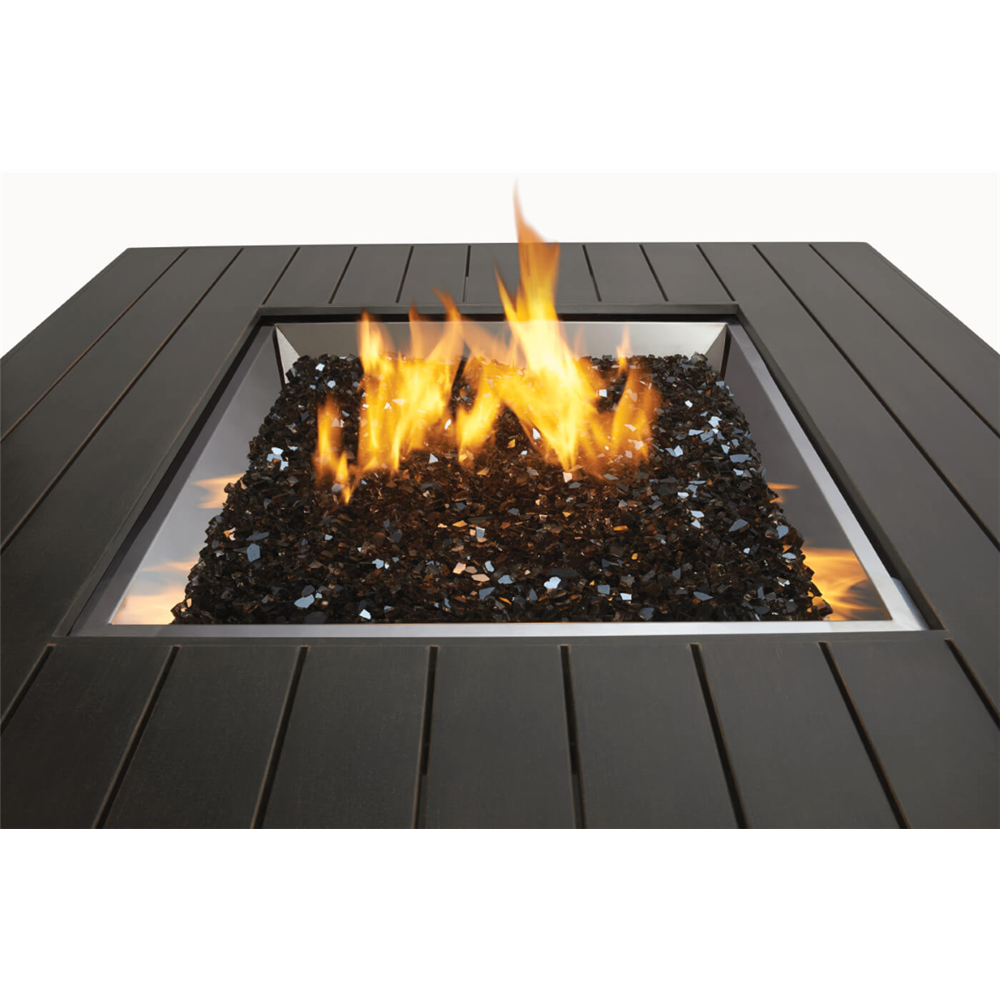 A square outdoor fire pit with a dark charcoal slatted surround, filled with black glass stones and active orange and yellow flames, shown against a plain white background.